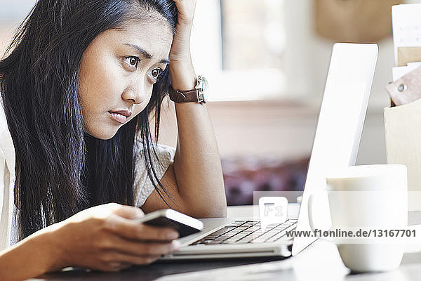 Unhappy young woman sitting at desk with laptop and mobile phone