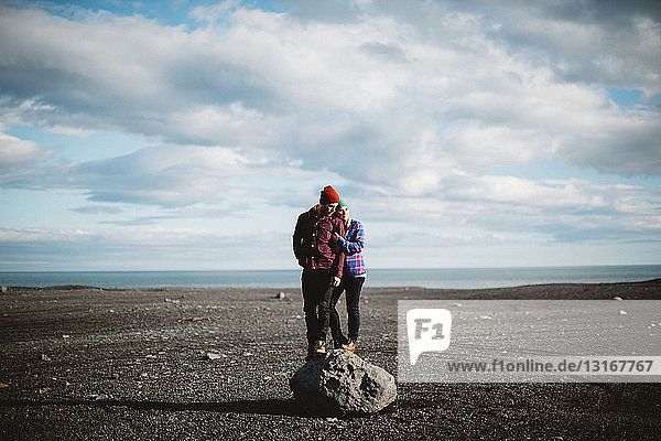 Mittelgroßes erwachsenes Paar auf einem Felsblock in vulkanischer Landschaft stehend  wegschauend  Island