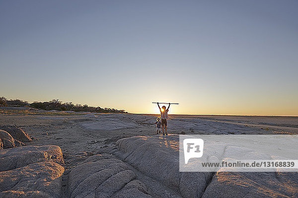 Zwei Jungen stehen auf einem Felsen und halten einen Speer in der Luft  Sonnenuntergang  Gweta  makgadikgadi  Botswana