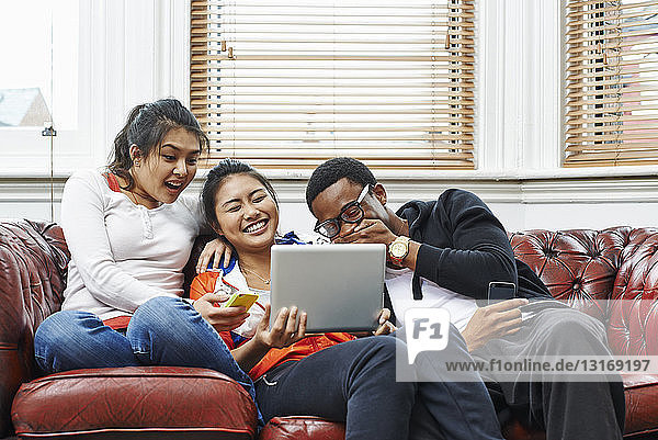 Drei junge Erwachsene sitzen auf dem Sofa und lachen über das digitale Tablet
