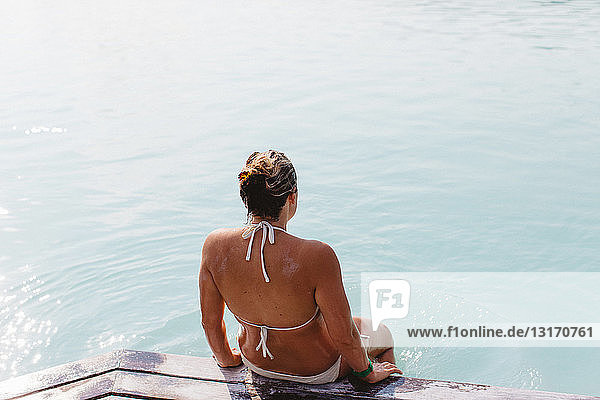 High angle rear view of mid adult woman wearing bikini sitting on pier  Blue Lagoon hot springs  Iceland