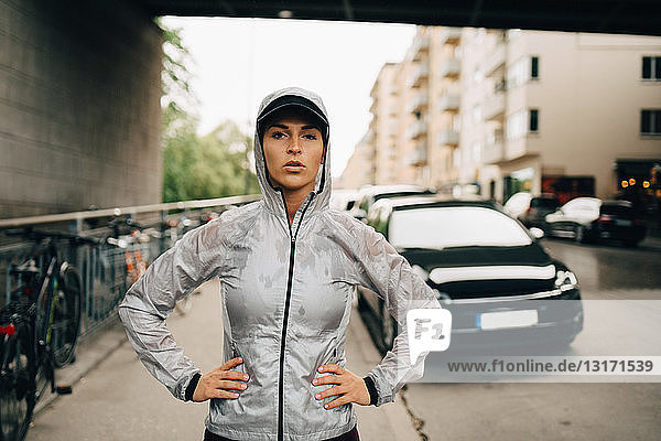 Portrait of confident female athlete in wet hooded jacket standing on sidewalk in city