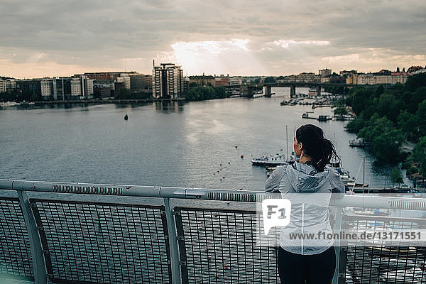 Rückansicht einer Athletin  die bei Sonnenuntergang auf einer Fußgängerbrücke über dem Meer steht und auf die Stadt schaut