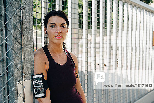 Portrait of confident sportswoman with smart phone and in-ear headphones leaning on railing at bridge