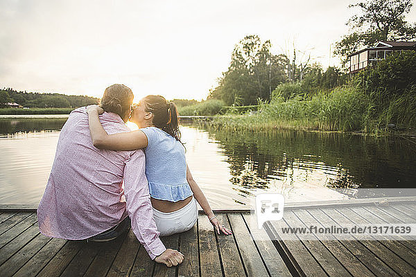 Rückansicht eines Freundes  der seine Freundin küsst und mit umarmten Armen auf dem Steg über dem See bei Sonnenuntergang sitzt