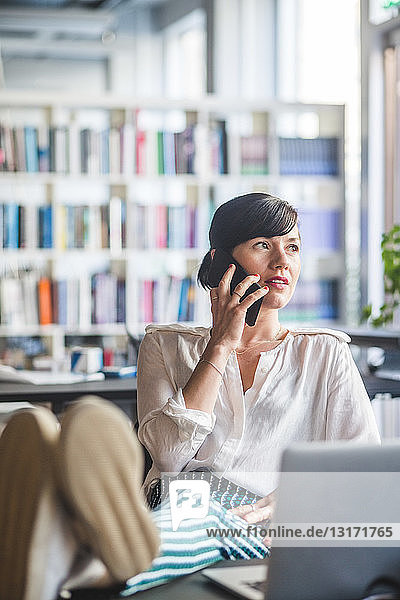 Creative businesswoman talking on mobile phone while relaxing at desk in office
