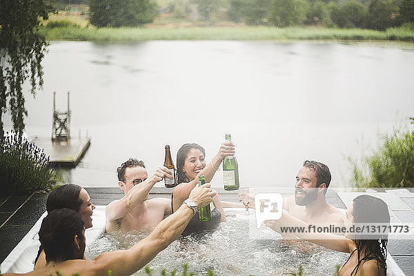 Carefree male and female friends enjoying drinks in hot tub against lake during weekend getaway
