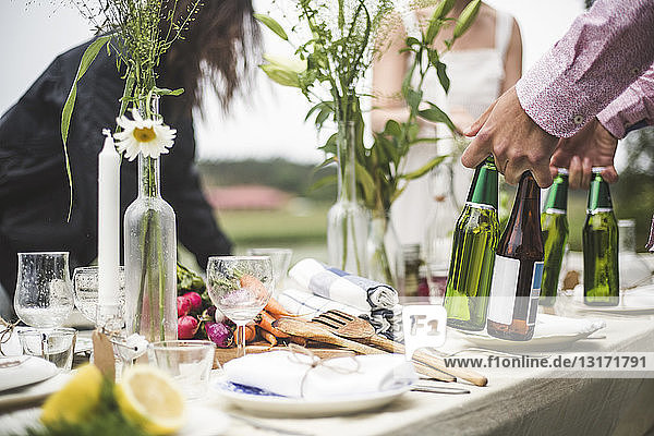 Cropped image of man holding beer bottles at dining table during dinner party in backyard