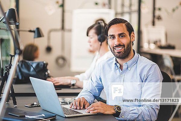 Portrait of confident businessman using laptop at desk in creative office