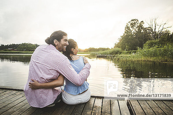Rückansicht eines lächelnden Paares  das bei Sonnenuntergang mit umgebundenen Armen auf dem Steg über dem See sitzt