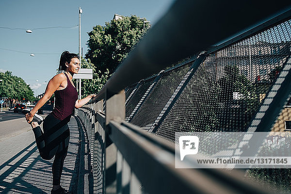 Junge Sportlerin streckt Bein am Geländer auf einer Brücke in der Stadt