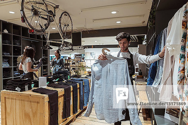 Salesman arranging clothes on rack while saleswoman assisting customers at store