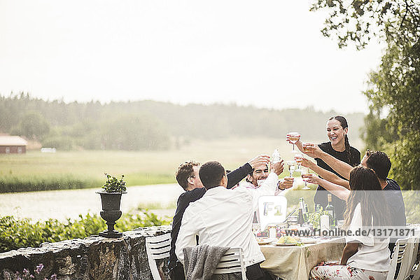 Fröhliche männliche und weibliche Freunde stoßen während der Dinnerparty im Hinterhof auf Weingläser an
