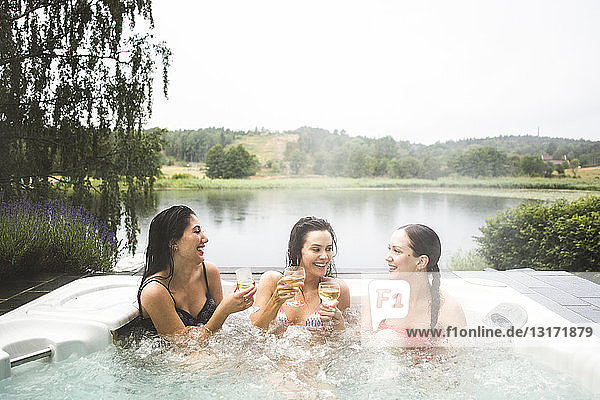 Cheerful female friends enjoying wine in hot tub against lake during weekend getaway