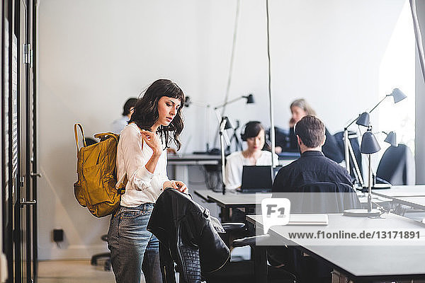 Creative businesswoman with backpack standing by chair in office
