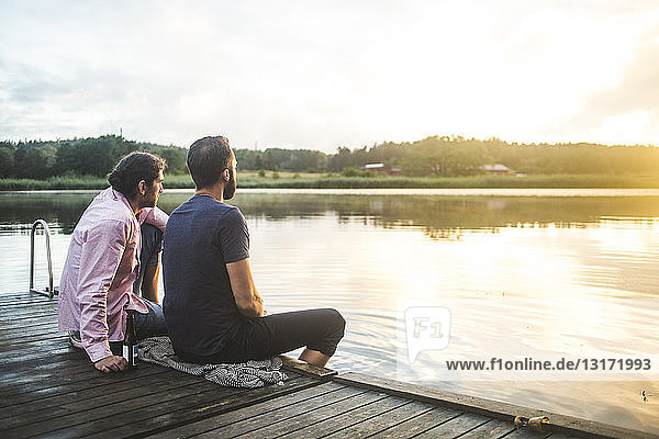Männliche Freunde schauen auf den See  während sie bei Sonnenuntergang auf dem Steg sitzen