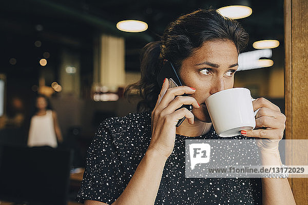 Mid adult businesswoman drinking coffee while using smart phone in office