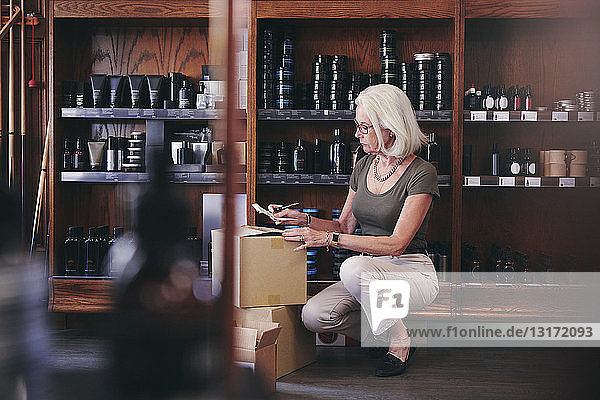 Leitende Verkäuferin hält Papier in der Hand  während sie im Feinkostladen an Kisten kauert