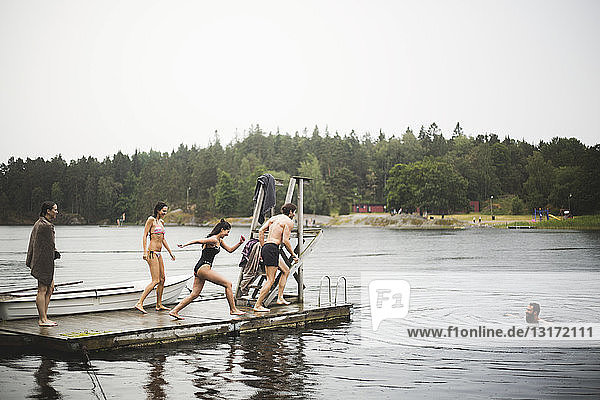 Sorglose männliche und weibliche Freunde rennen auf einem Steg zum Tauchen im See während eines Wochenendausflugs