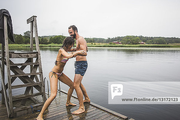 Verspielte Frau schiebt einen männlichen Freund in den See  während sie während eines Wochenendausflugs auf einem Steg steht