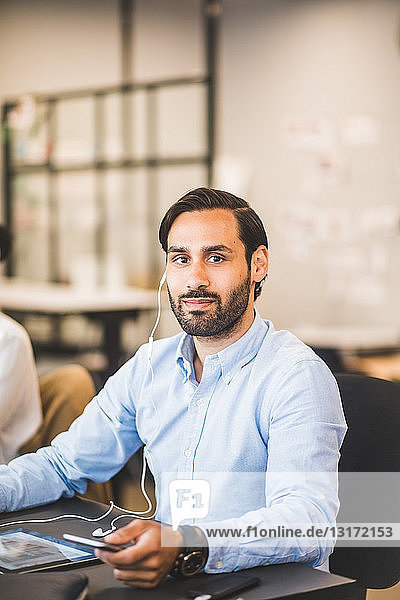 Portrait of confident businessman using in-ear headphones while sitting at desk in office