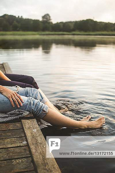Low section of female friends dangling legs in lake while sitting on jetty