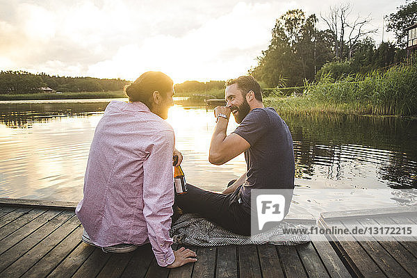 Smiling male friends talking while sitting on jetty over lake during sunset