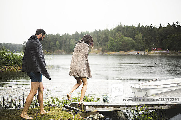 In Handtücher gewickelte Freundinnen und Freunde gehen während eines Wochenendausflugs zum Steg über dem See