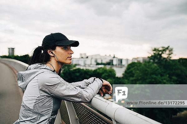 Tired sportswoman looking away while leaning on railing at footbridge in city