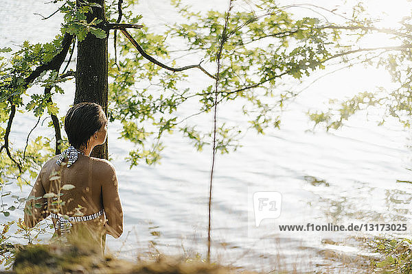 Rückansicht einer Frau im Bikini am Baumstamm an einem See