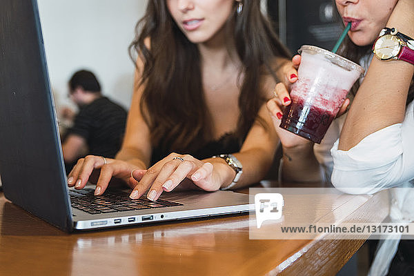 Young woman drinking smoothie in a coffee shop while her friend using laptop  partial view