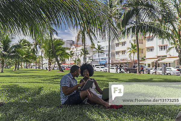 USA  Florida  Miami Beach  young couple using tablet and laptop on lawn in a park