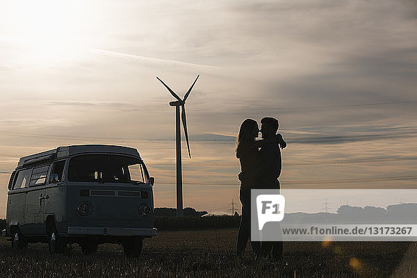 Young couple kissing at camper van at dusk with wind turbine in background