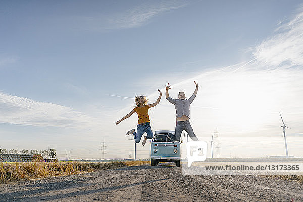 Exuberant couple jumping on dirt track at camper van in rural landscape