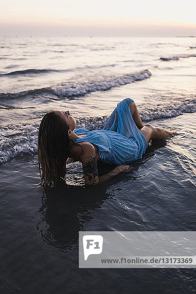 Junge tätowierte Frau in blauem Kleid liegt bei Sonnenuntergang im Wasser am Meeresufer