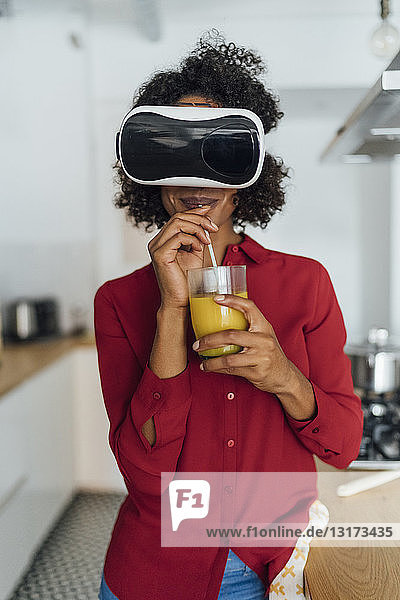 Woman standing in her kitchen  wearing VR goggles  drinking orange juice