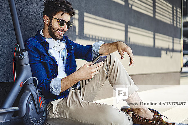 Lächelnder junger Mann sitzt neben seinem Elektroroller auf dem Boden und benutzt sein Handy