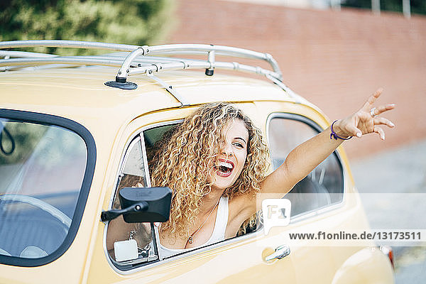 Portrait of happy blond woman leaning out of window of classic car showing victory sign