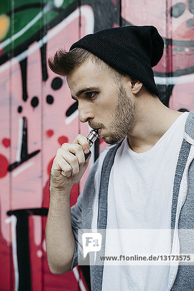 Junger Mann steht vor Graffiti und raucht elektronische Zigarette