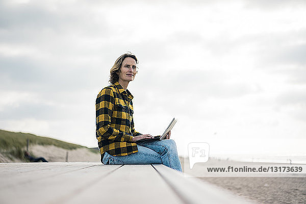 Reife Frau sitzt am Strand auf der Strandpromenade und benutzt einen Laptop