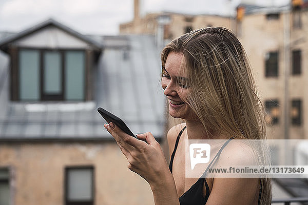 Lächelnde blonde junge Frau auf der Dachterrasse  die auf ihr Handy schaut