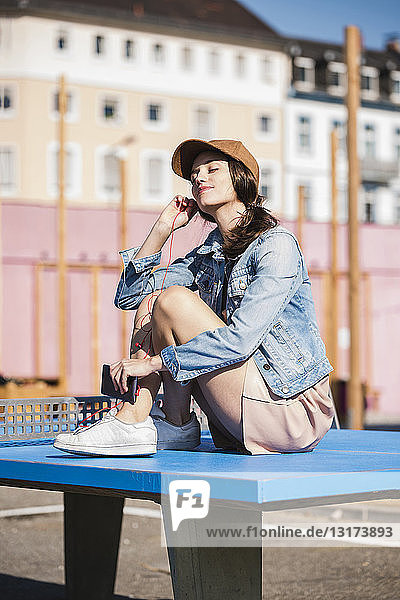 Entspannte junge Frau sitzt auf einer Tischtennisplatte und hört Musik