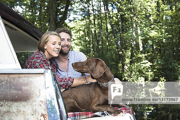 Smiling young couple with dog sitting in car at a brook in forest