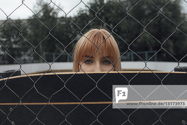 Porträt einer jungen Frau hinter Carver-Skateboard und Maschendrahtzaun