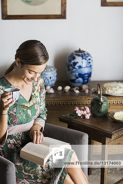 Young woman drinking cup of tea while reading a book at home