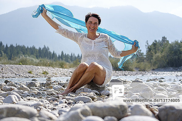 Glückliche Frau hält schwingenden blauen Schal am Flussufer  Oberbayern  Deutschland