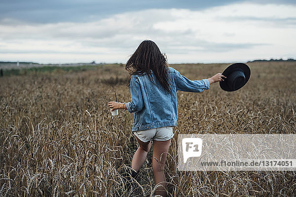 Back view of young woman with beverage and hat walking in corn field