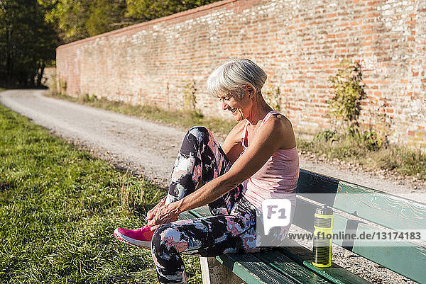 Lächelnde sportliche ältere Frau sitzt auf einer Bank und bindet sich die Schuhe