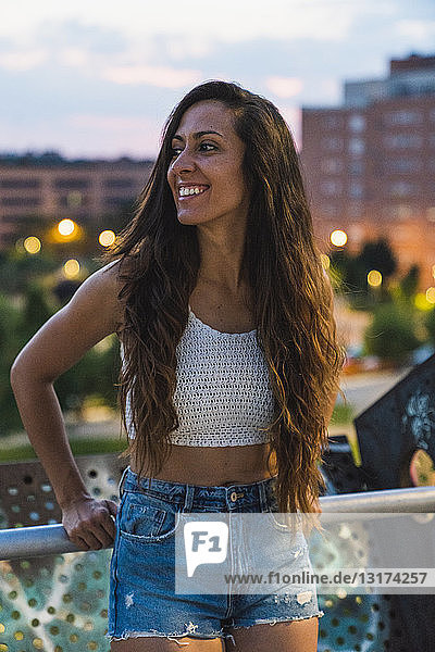 Schöne lächelnde junge Frau mit langen braunen Haaren in der Stadt in der Abenddämmerung