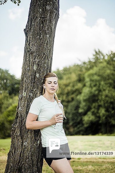 Sportliche junge Frau  die sich in einem Park mit einer Flasche an einen Baum lehnt